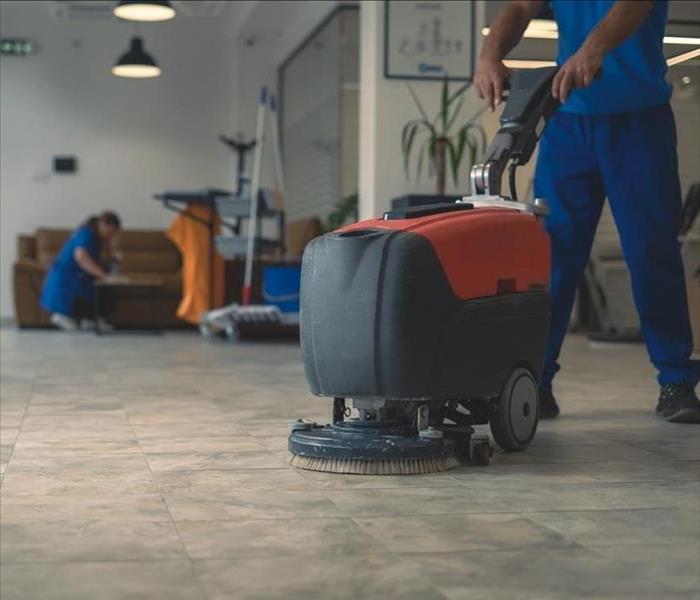 Cleaner cleans hard floor with scrubber machine while other cleaner cleans in the background