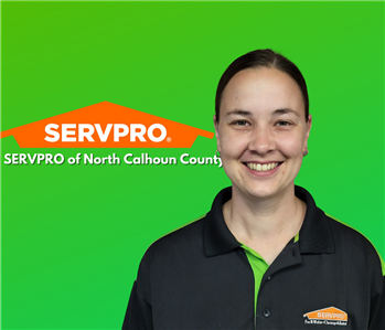 Caucasian young female in black and green shirt, burnette hair pulled back. Standing against green wall with SERVPRO logo.