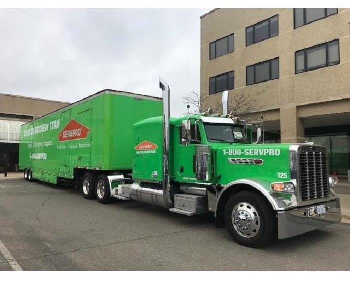 Large green SERVPRO semi in front of large beige building at water loss, with trees surrounding it.